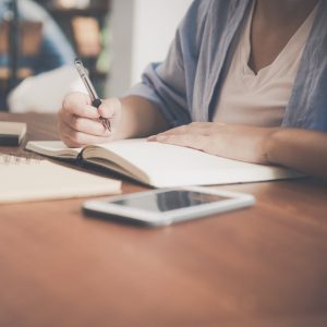 woman writing on a notebook beside teacup and tablet computer